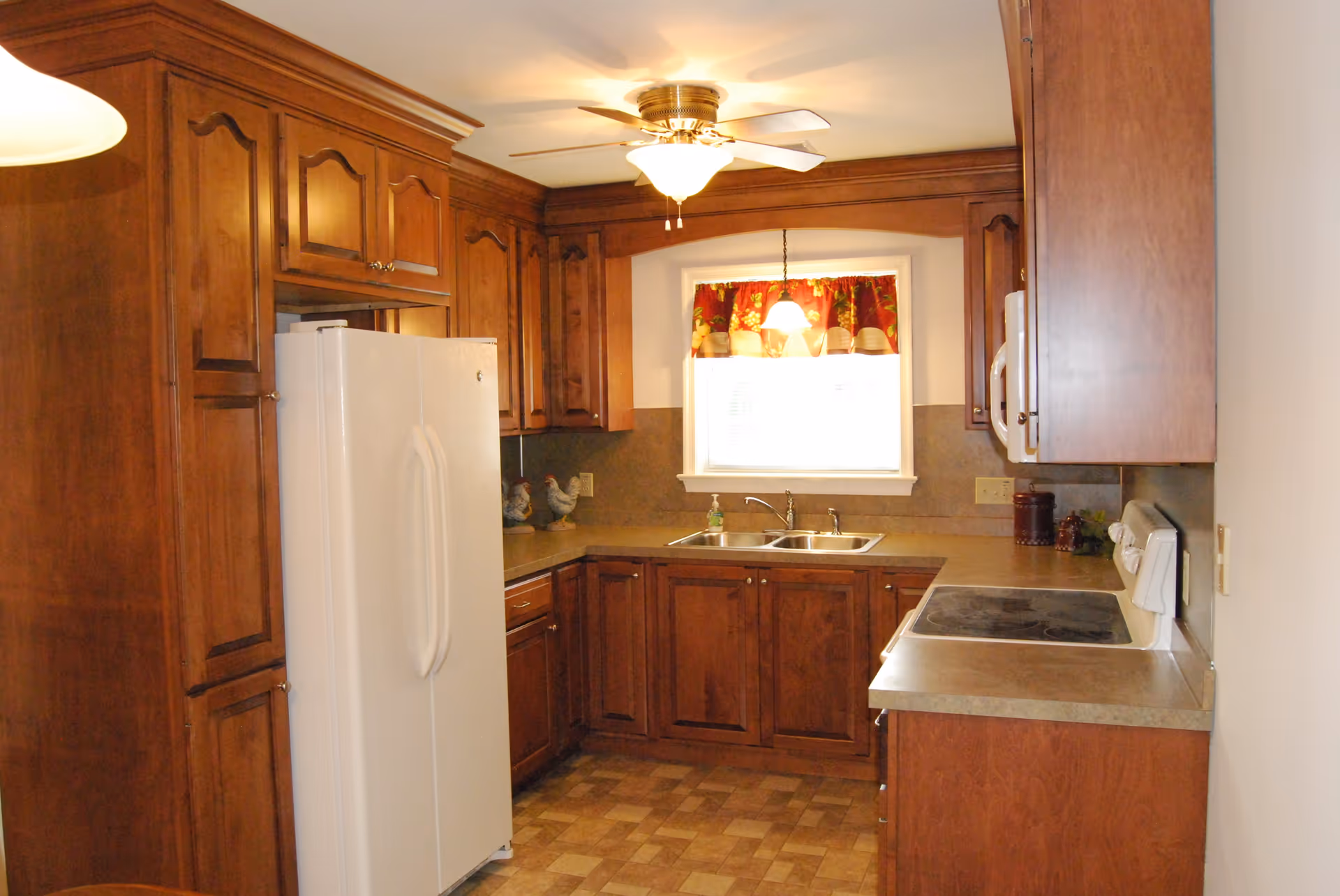Well-lit kitchen with wooden cabinets, a white refrigerator, double sink beneath a curtained window, and an electric stovetop.
