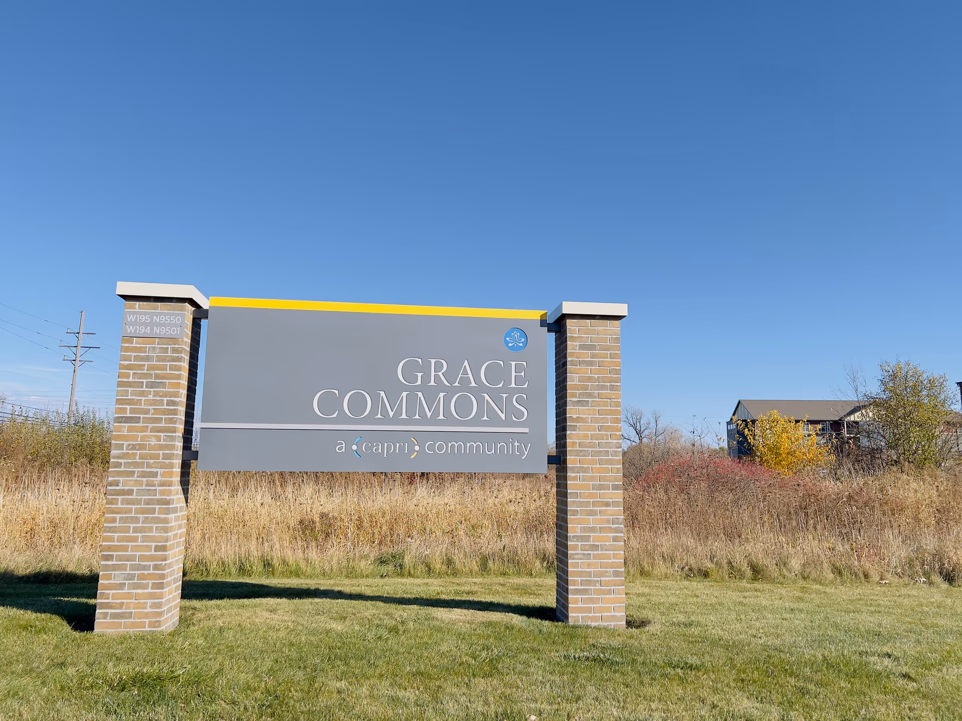 Outdoor sign for Grace Commons, a Capri community, mounted on two brick pillars with grass and shrubs in the background under a clear blue sky.