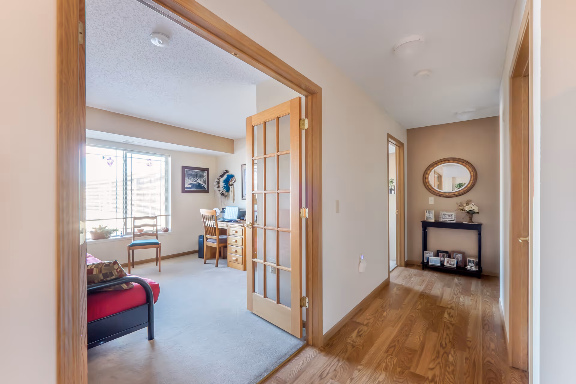 View of a hallway with wooden flooring leading to a room with a desk, chair, and window. The room has a red cushioned bench, a wooden chair, and framed pictures on the walls. A wooden-framed glass door is open between the hallway and the room. The hallway has a small black table with framed photos and a round mirror above it.