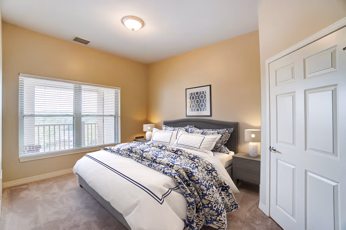 A bedroom with a large bed covered in white and blue patterned bedding, two bedside tables each with a lamp, a framed abstract artwork on the wall above the bed, a large window with blinds letting in natural light, beige walls, and a closed white door.