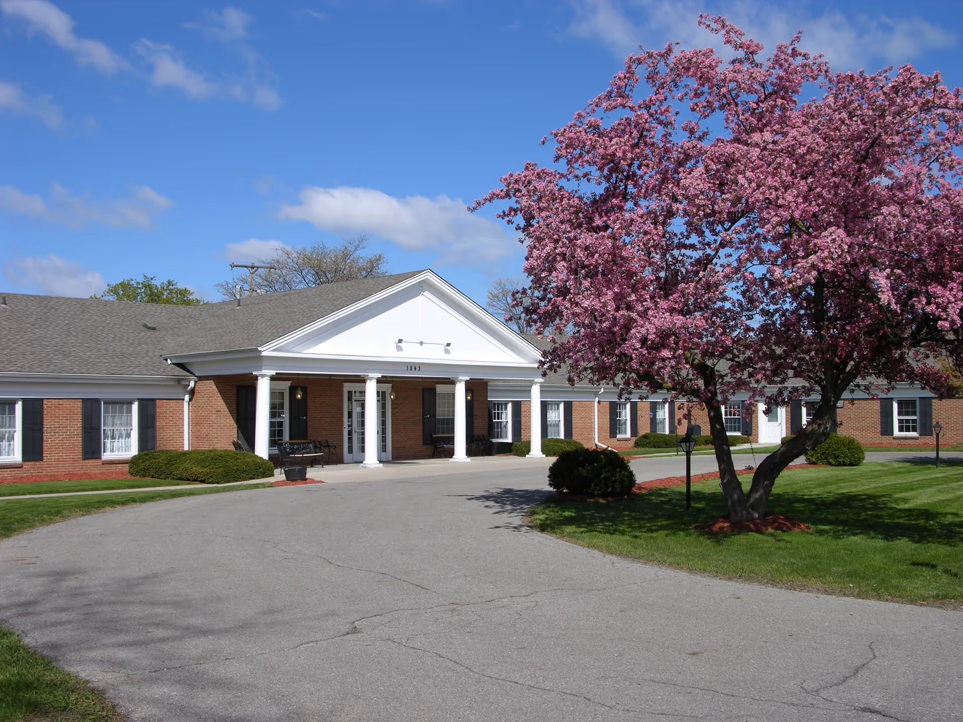 Single-story brick senior living facility entrance with white columns and a blooming pink tree in the front lawn.