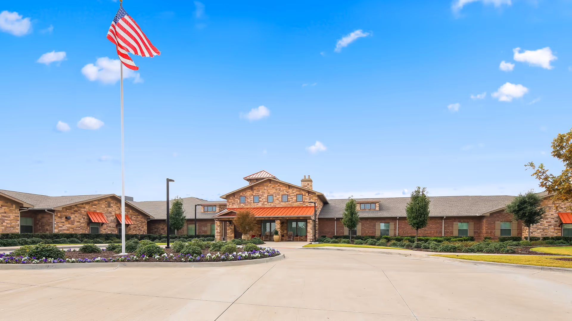 Front exterior view of Rock Ridge Assisted Living and Memory Care facility with a large American flag on a flagpole, a circular driveway, landscaped flower beds, and a clear blue sky with scattered clouds.