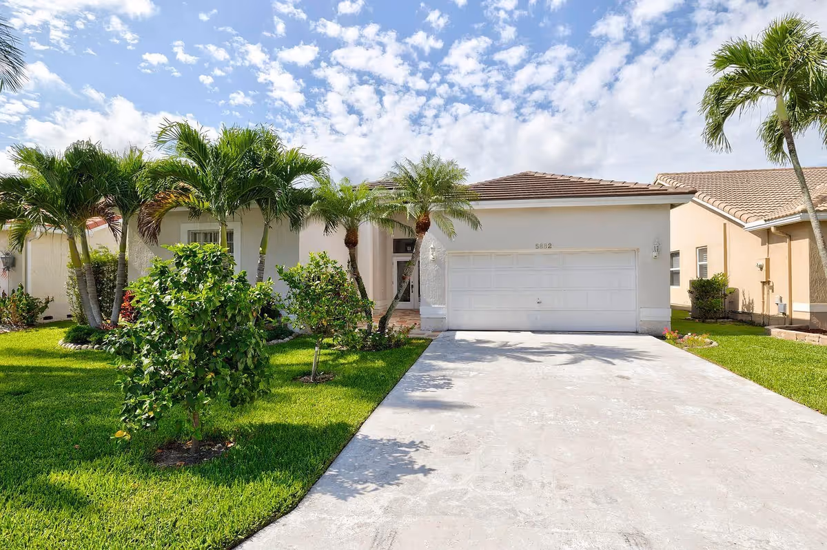 Front exterior view of a single-story house with a two-car garage, a concrete driveway, and a well-maintained lawn with palm trees and shrubs under a partly cloudy sky.