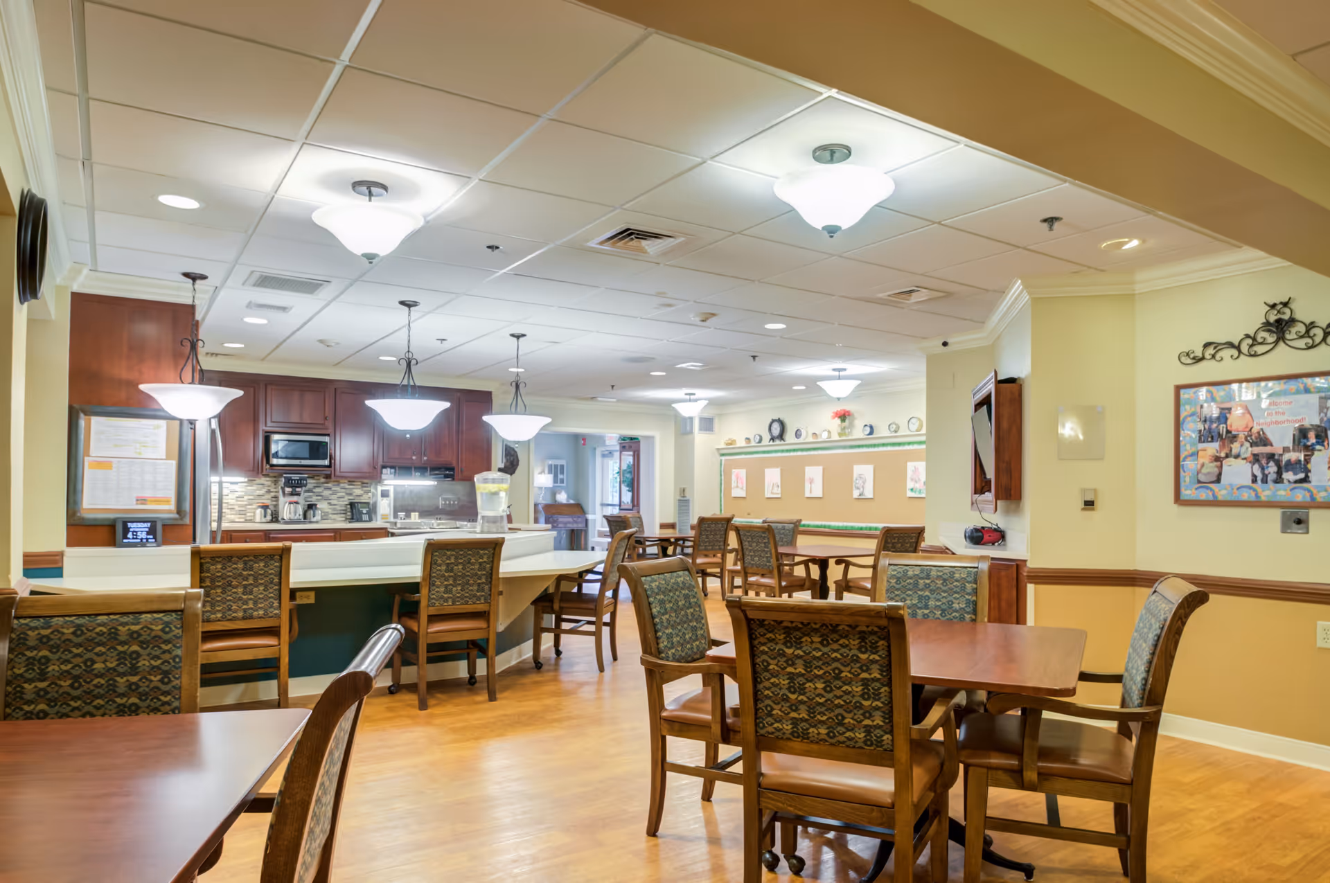 A well-lit dining area in a senior living facility featuring wooden tables and chairs with patterned upholstery. The room includes a kitchen area with dark wood cabinets, a microwave, and a coffee maker. Pendant lights hang over the kitchen counter, and the walls are decorated with framed pictures and a bulletin board. The floor is wooden, and the ceiling has recessed lighting and ceiling tiles.