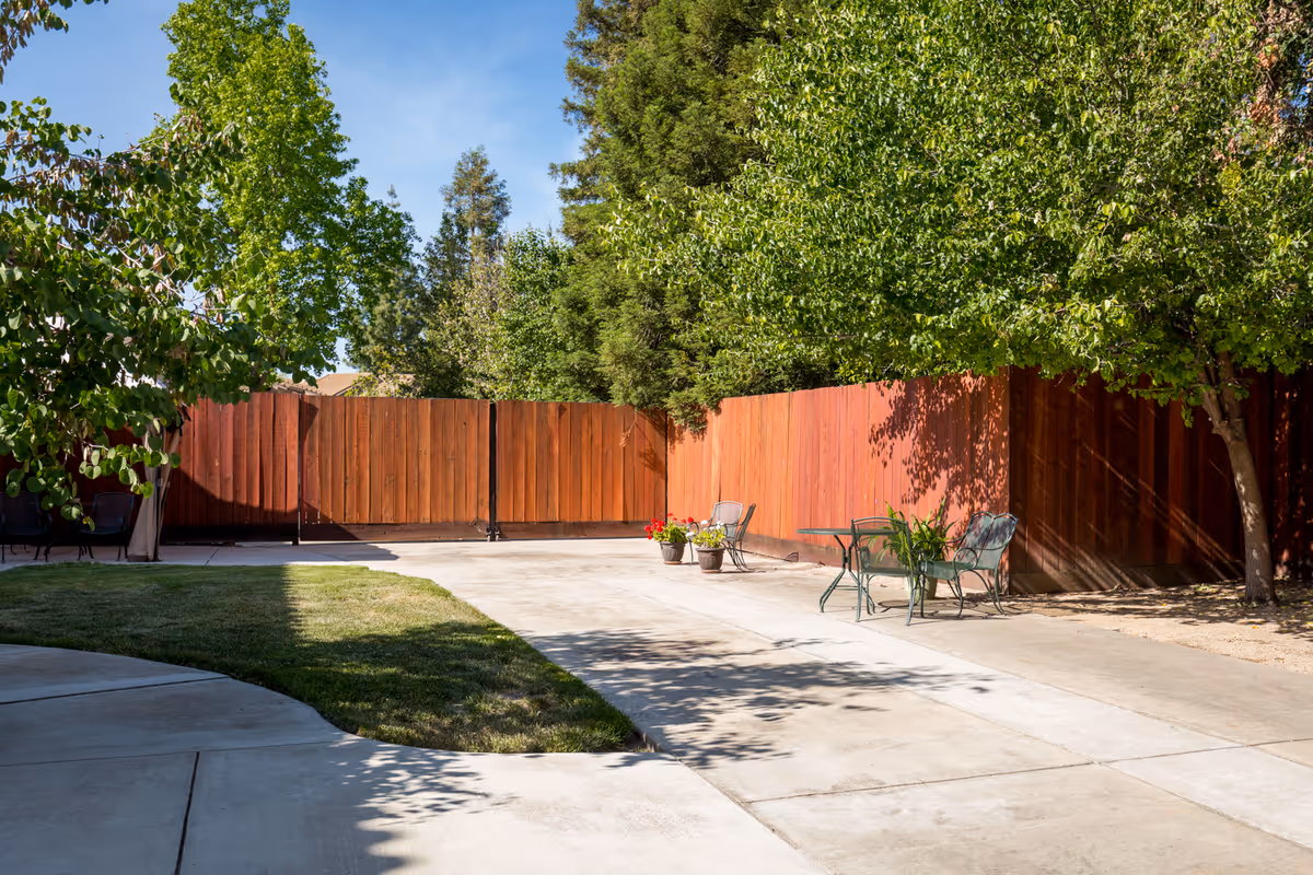 Outdoor patio area with concrete pavement, green grass, and several trees. There are two metal chairs and a small table with a plant on it, as well as two potted plants with flowers near a wooden fence. The sky is clear and blue.