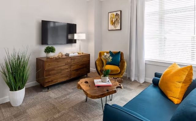 A cozy living room with a blue sofa adorned with a yellow pillow, a unique wooden coffee table with a small plant and books, a mustard yellow armchair with a teal pillow, a wooden media console with a potted plant and lamp, a wall-mounted flat-screen TV, a framed abstract artwork, and a large window with white blinds and light gray curtains.