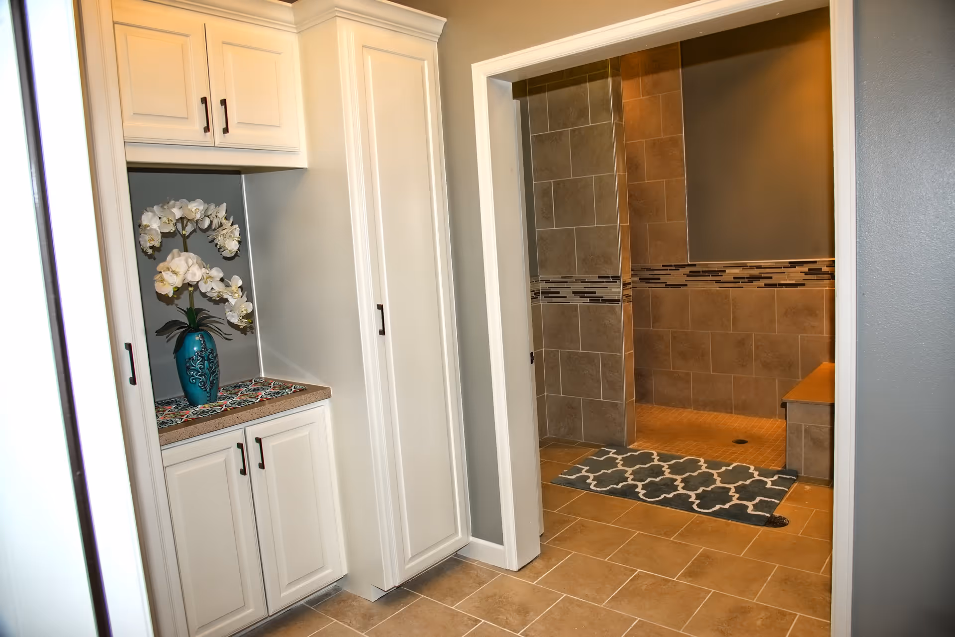 Interior view of a tiled bathroom area with a walk-in shower featuring beige tiles and a decorative horizontal tile strip. Adjacent to the shower is a built-in cabinet with white doors and black handles, topped with a countertop holding a blue vase with white flowers. The floor is tiled in a matching beige color, and a patterned rug is placed in front of the shower entrance.