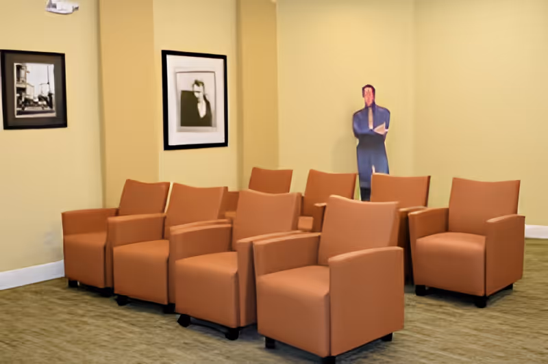 A room with eight orange armchairs arranged in two rows facing forward, beige walls with two framed black and white pictures, and a life-sized cardboard cutout of a person standing in the back corner.