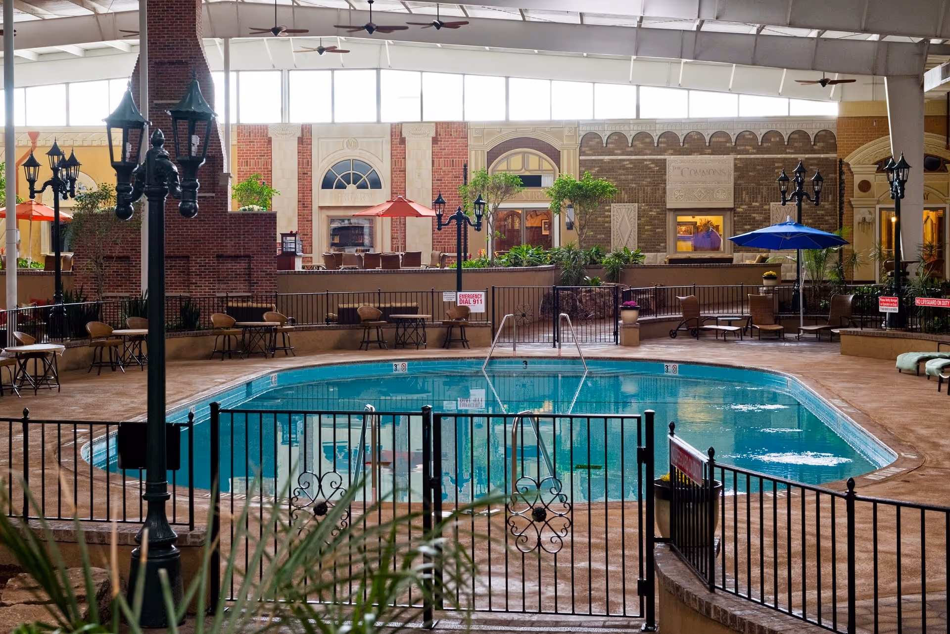 Indoor swimming pool area with a black metal fence surrounding the pool. Several lounge chairs and tables with umbrellas are placed around the pool. The background features a brick wall with arched windows and decorative elements, and ceiling fans hang from the high ceiling.