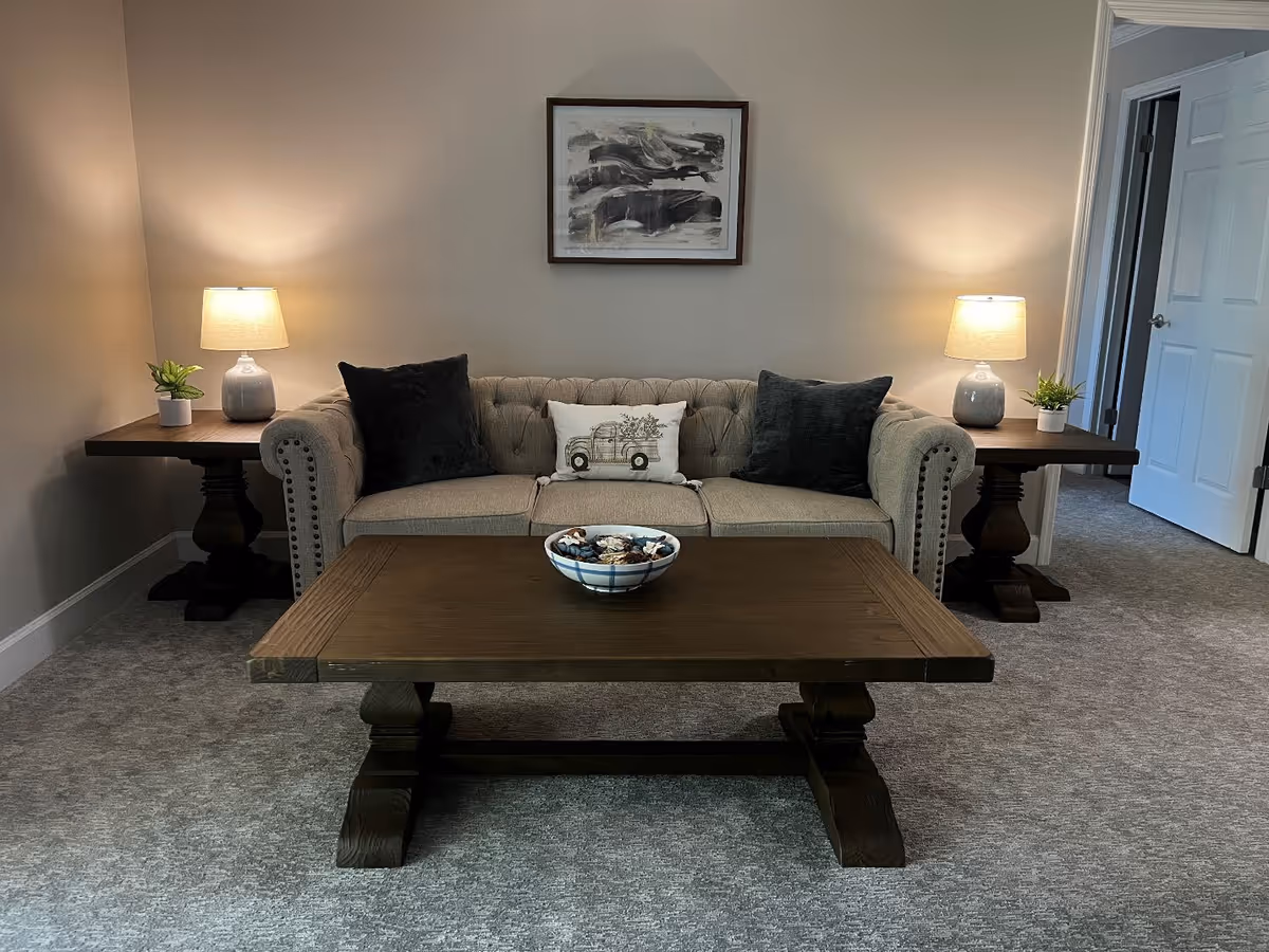 A cozy living room with a beige tufted sofa adorned with two dark gray pillows and a decorative pillow featuring a vintage truck. On either side of the sofa are dark wooden side tables with matching lamps and small potted plants. A wooden coffee table with a decorative bowl sits in front of the sofa. The walls are light-colored with a framed abstract painting hanging above the sofa. A doorway is visible to the right.