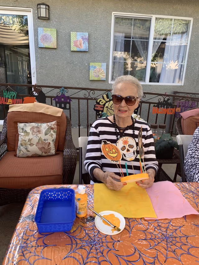 An older woman wearing sunglasses sits at a Halloween-decorated outdoor table holding pumpkin and skull craft decorations.