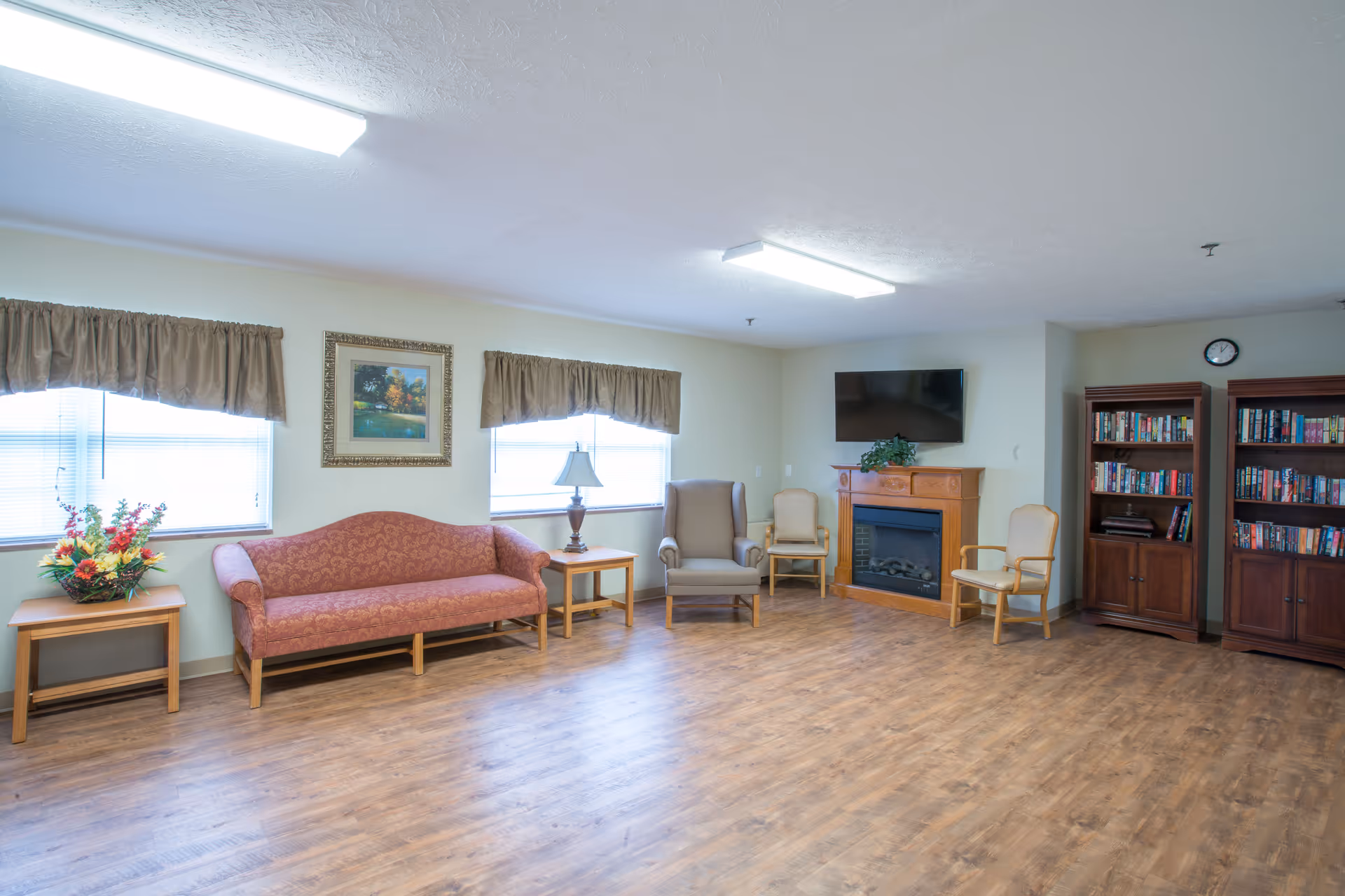A bright and spacious common area with wooden flooring, featuring a red patterned sofa, two beige armchairs, a wooden side table with a lamp, a fireplace with a TV mounted above it, two wooden bookshelves filled with books, and two windows with brown valances letting in natural light.