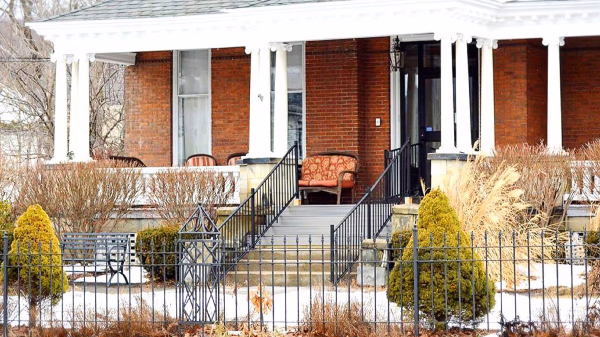 Front of a brick building showing a covered porch with chairs, steps and a black iron fence surrounded by snow and landscaping.