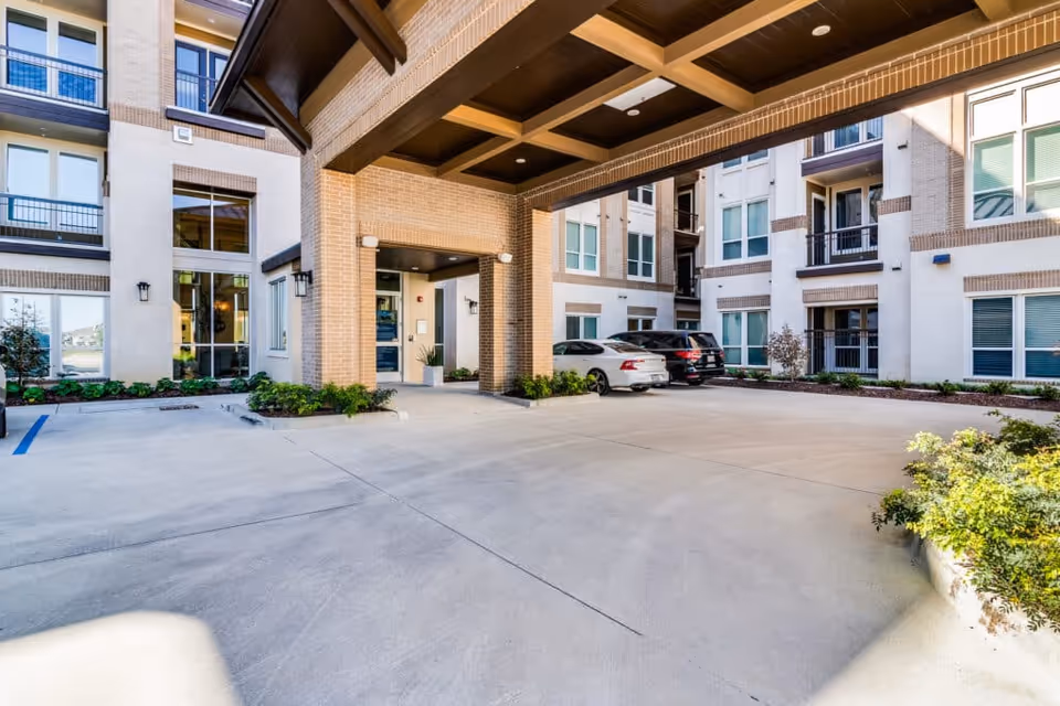 Exterior view of the entrance area of The Villages Of Imperial Pointe, showing a covered driveway with parked cars and surrounding multi-story building with windows and balconies.