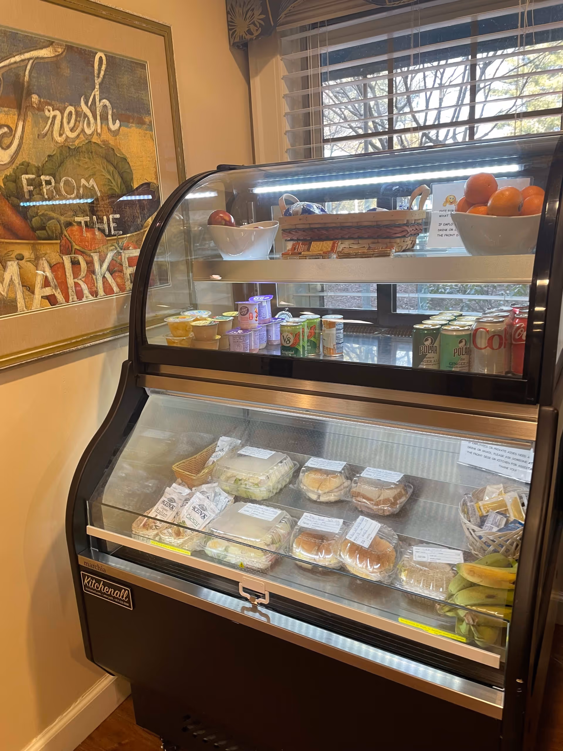 A refrigerated display case in a dining area containing packaged sandwiches, yogurt cups, canned beverages, and bowls of fresh fruit. A window with blinds and a framed artwork with the text 'Fresh from the Market' are visible in the background.