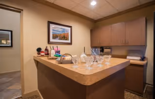 Interior view of a small kitchen or serving area with a countertop displaying several empty glass cups, a basket with bread, and some condiments. There are wooden cabinets on the wall and a framed landscape picture hanging above the counter.