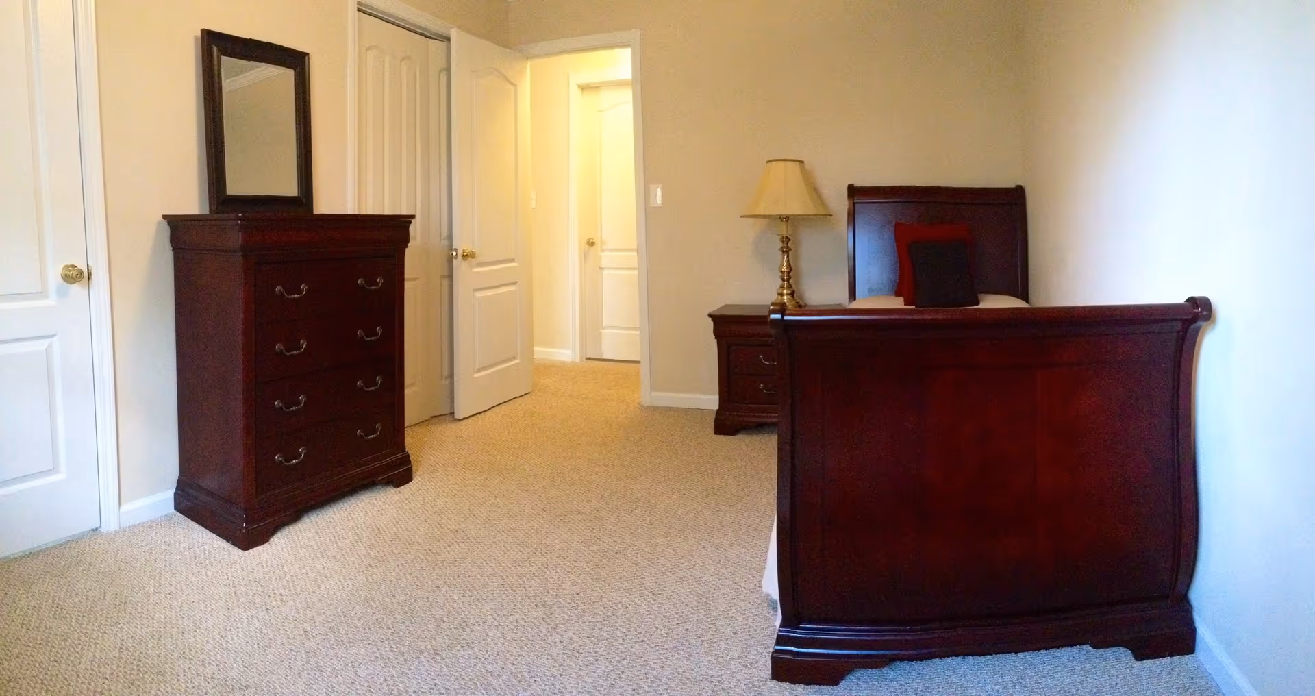 A bedroom with beige walls and carpeted floor featuring a dark wooden bed with a red and black pillow, a matching nightstand with a lamp, and a tall dresser with a mirror. Two white doors are visible, one open leading to a hallway.