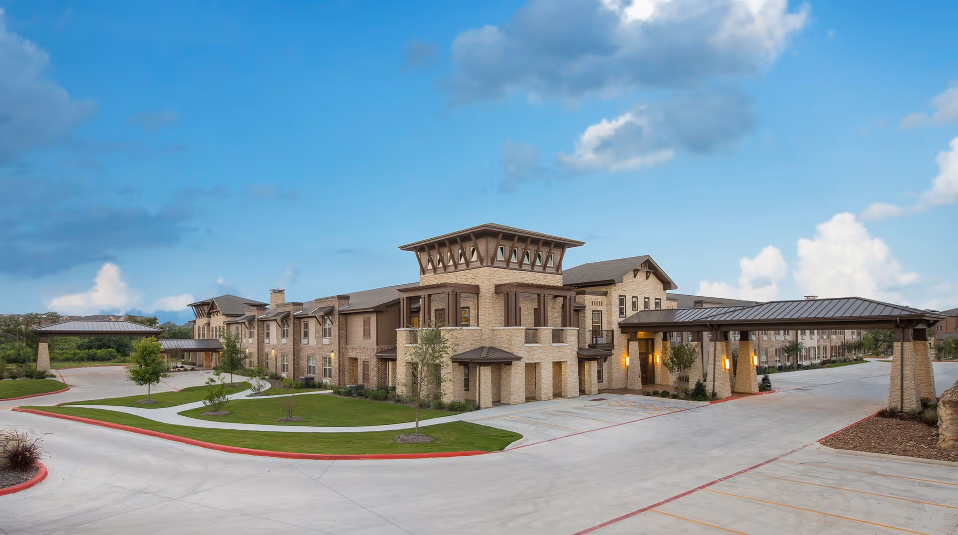 Exterior view of a large, modern assisted living and memory care facility building with stone and wood accents, surrounded by landscaped green lawns and a paved driveway under a partly cloudy blue sky.