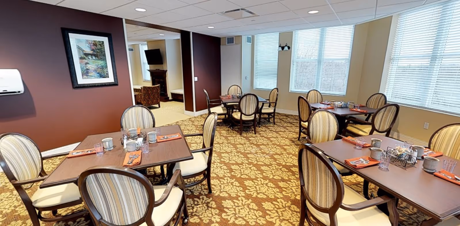 A dining room with several wooden tables and cushioned chairs arranged neatly. Each table is set with cups, glasses, napkins, and silverware. The room has large windows with blinds allowing natural light to fill the space. The walls are painted in a combination of maroon and beige, and there is a framed painting on one wall. The carpet has a floral pattern in shades of brown and beige.