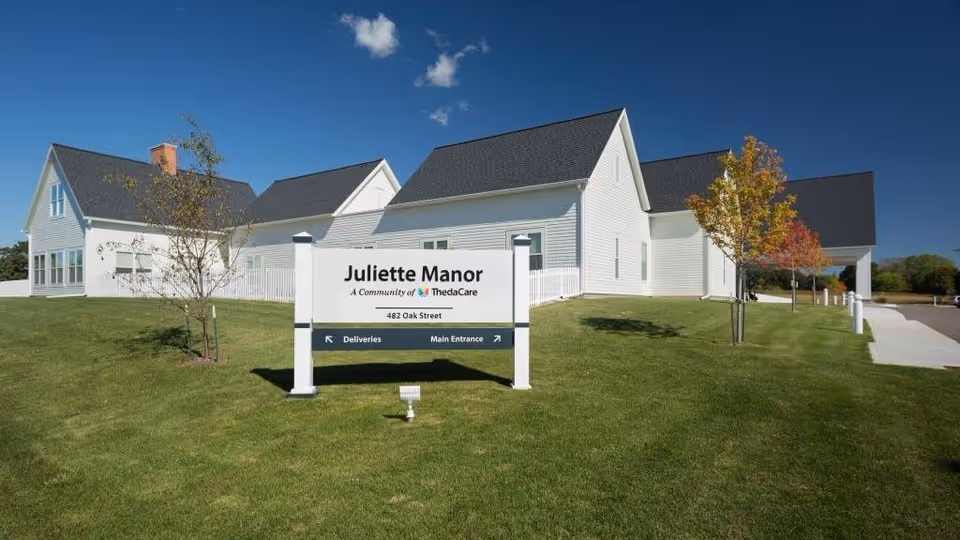 Exterior view of Juliette Manor, a white building with dark roof shingles, situated on a well-maintained grassy lawn with a few small trees. A sign in front reads 'Juliette Manor, A Community of ThedaCare, 482 Oak Street' with directional arrows for Deliveries and Main Entrance. The sky is clear and blue with a few small clouds.