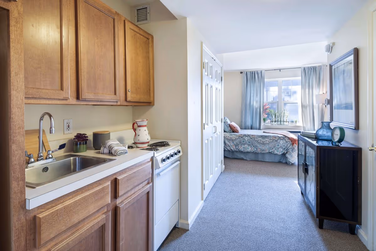 A small studio apartment with a kitchenette on the left featuring wooden cabinets, a sink, and a stove. The room extends to a sleeping area with a bed covered in patterned bedding, a window with light blue curtains, and a small cabinet with decorative items including a clock and a vase.