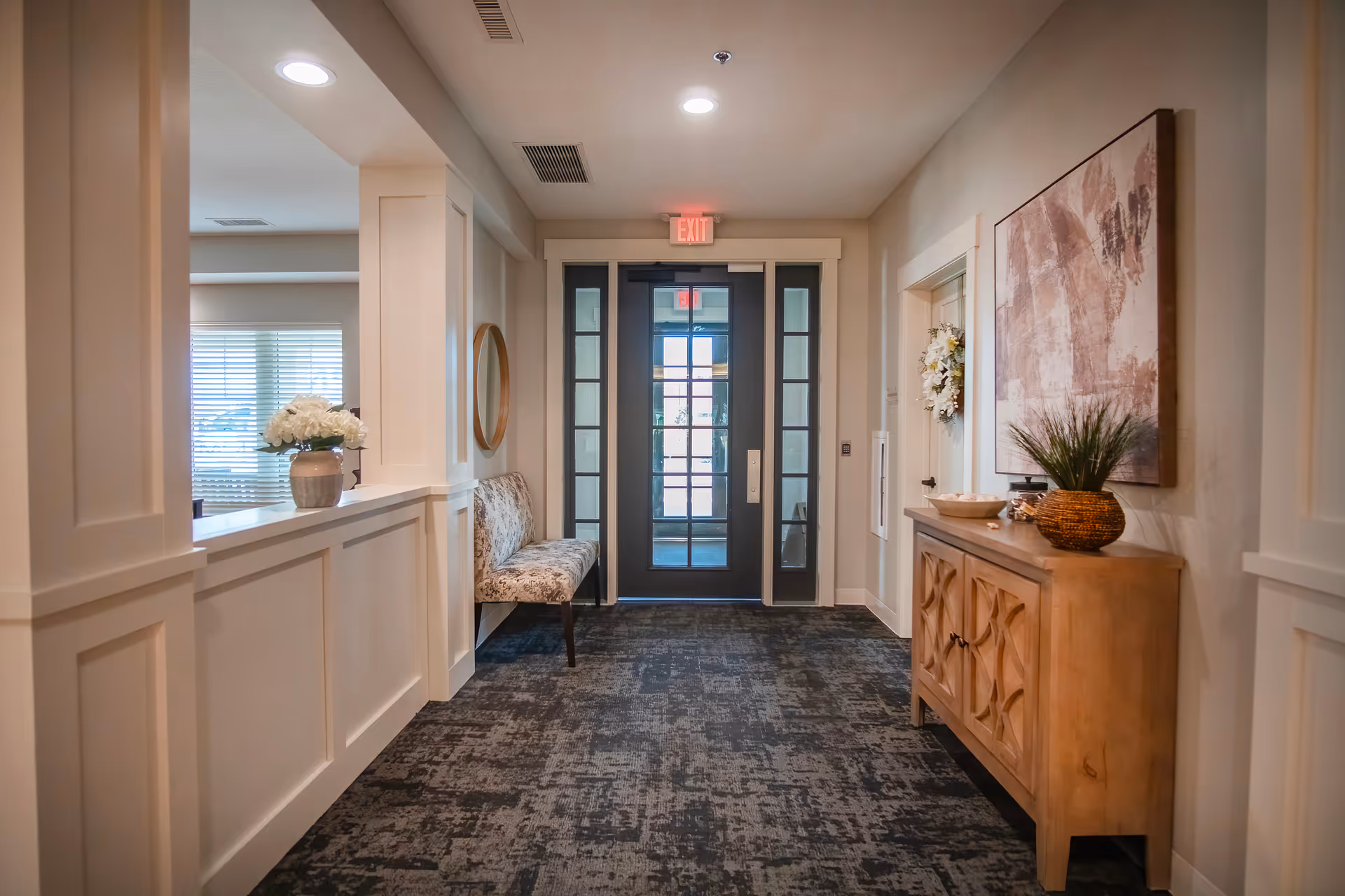 A well-lit hallway with a patterned carpet leading to a glass door with an exit sign above it. On the left side, there is a built-in bench with floral upholstery and a round mirror above it. On the right side, there is a wooden cabinet with decorative carvings, topped with a plant and a bowl, and a large abstract painting hanging above it. The walls are painted in a neutral tone, and there is a window with blinds visible through an opening on the left.
