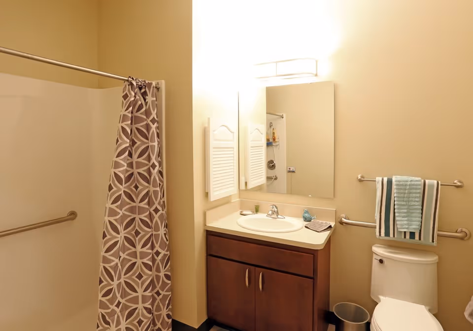 Well-lit bathroom showing a shower with patterned curtain, sink vanity and mirror, and a toilet with towel rack.