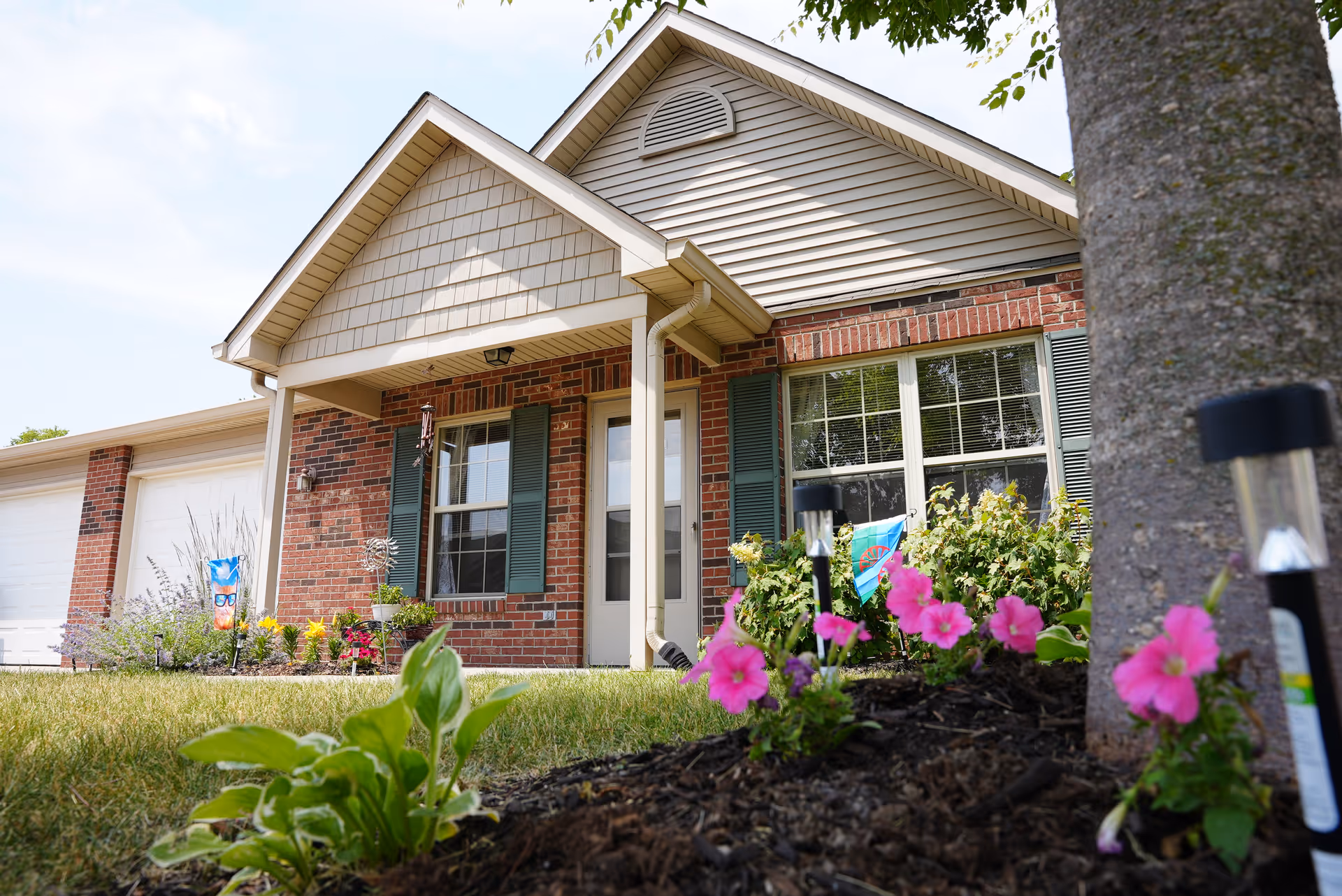 Exterior view of a single-story brick and siding house with a front porch, green shutters, a white door, and a garden bed with pink flowers and solar garden lights in the foreground.
