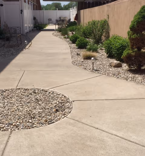 A paved outdoor walkway with a circular gravel feature in the foreground, bordered by gravel beds with various green shrubs and small plants. There are patio chairs and tables visible in the background near a white fence.