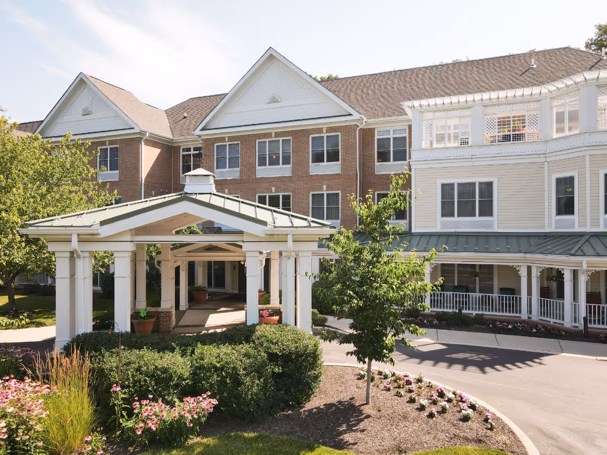 Exterior view of a senior living facility building with a covered entrance, surrounded by landscaped greenery and flowers under a clear sky.