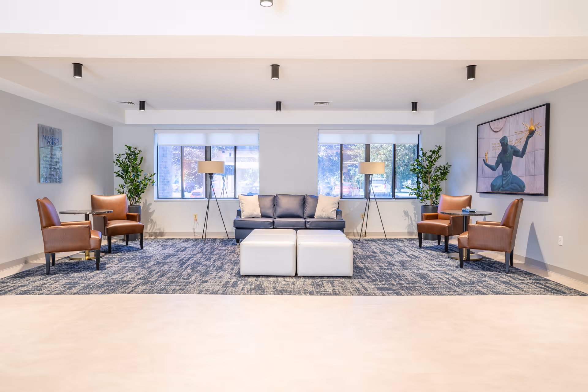 A bright and spacious lounge area with a blue sofa in the center, flanked by two white ottomans. On either side of the sofa are two brown leather armchairs with small round tables next to them. Two tall floor lamps stand behind the sofa near large windows letting in natural light. Green potted plants are placed near the armchairs. The walls are light-colored with a framed artwork on the right wall and a sign on the left wall. The floor has a blue patterned carpet in the seating area and light-colored flooring in the foreground.