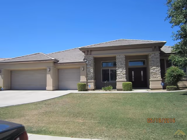 Front exterior view of a single-story residential building with a three-car garage, stone and stucco facade, a well-maintained lawn, and clear blue sky.