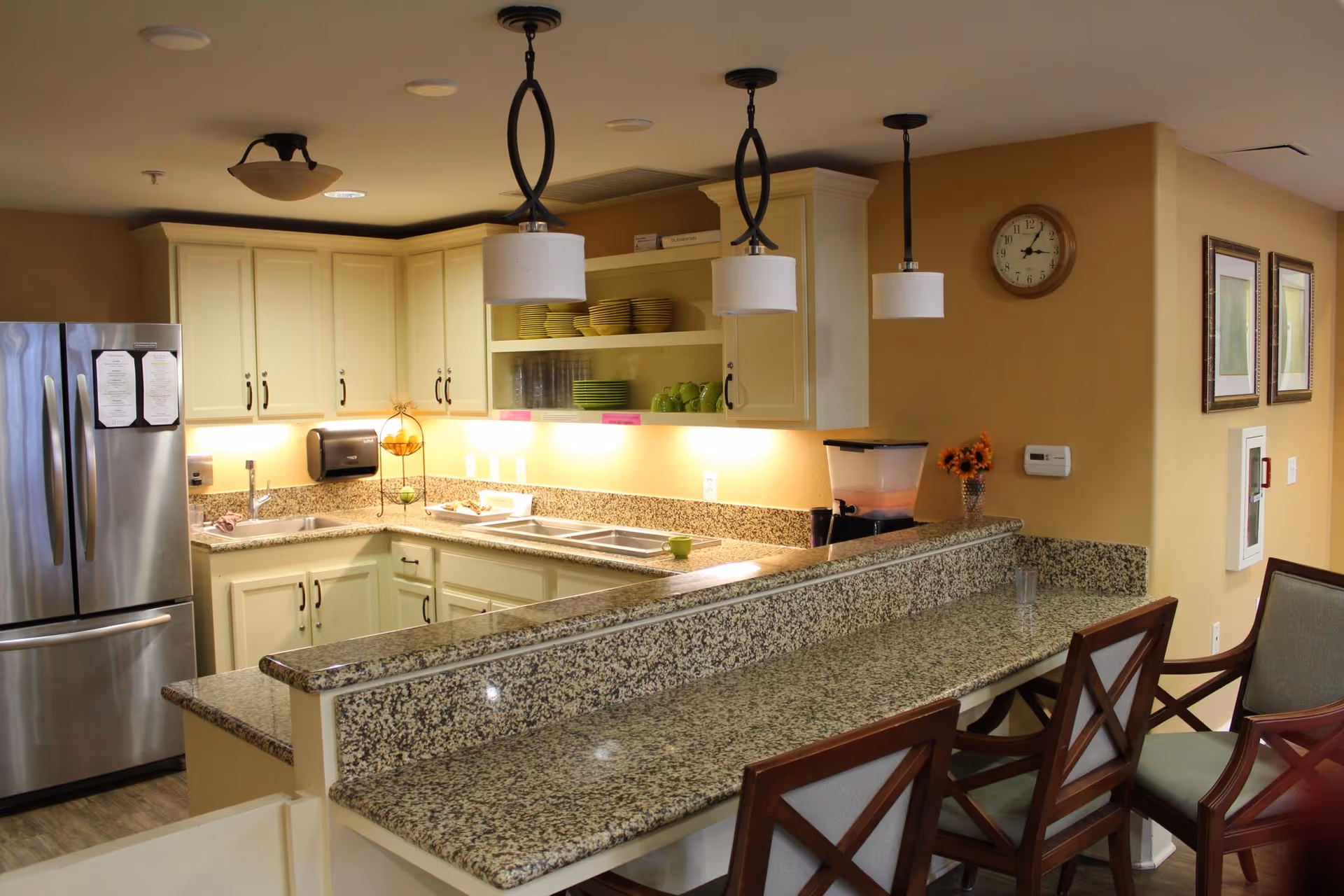 A clean and well-lit kitchen area with cream-colored cabinets, granite countertops, and a stainless steel refrigerator. There are three pendant lights hanging above the counter, which has three wooden chairs with cushioned seats. The kitchen includes a sink, open shelves with plates and cups, a paper towel dispenser, and a clock on the wall. The walls are painted a warm beige color.