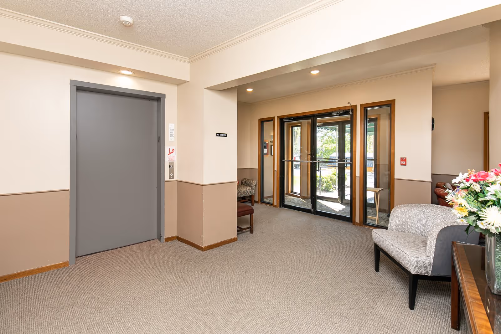 Interior view of a senior apartment building lobby showing a closed gray elevator door on the left, a seating area with chairs and a table with a vase of flowers on the right, and glass double doors leading outside in the background.