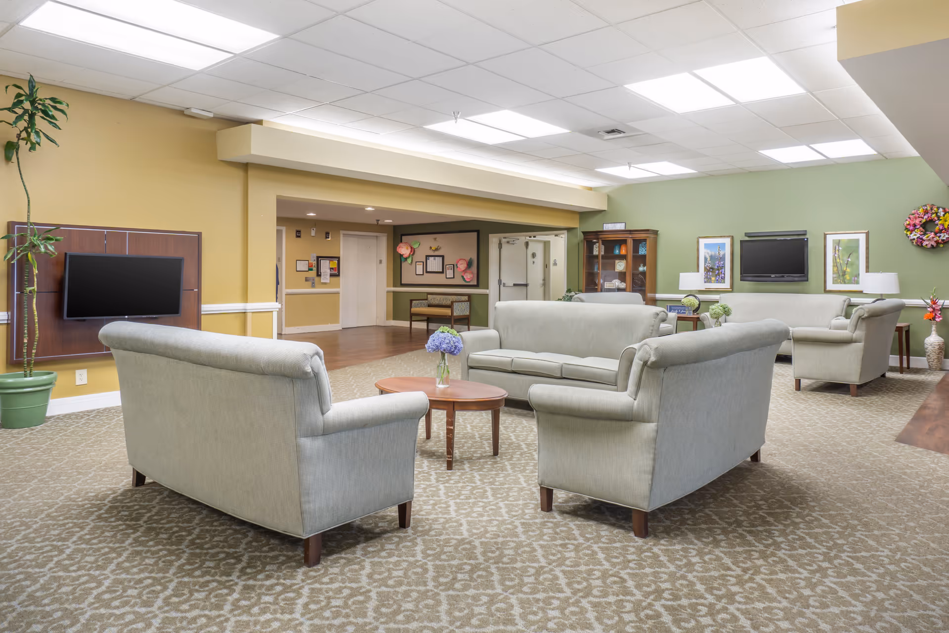 A spacious senior living lounge with multiple sofas, coffee tables, and a wall-mounted TV.