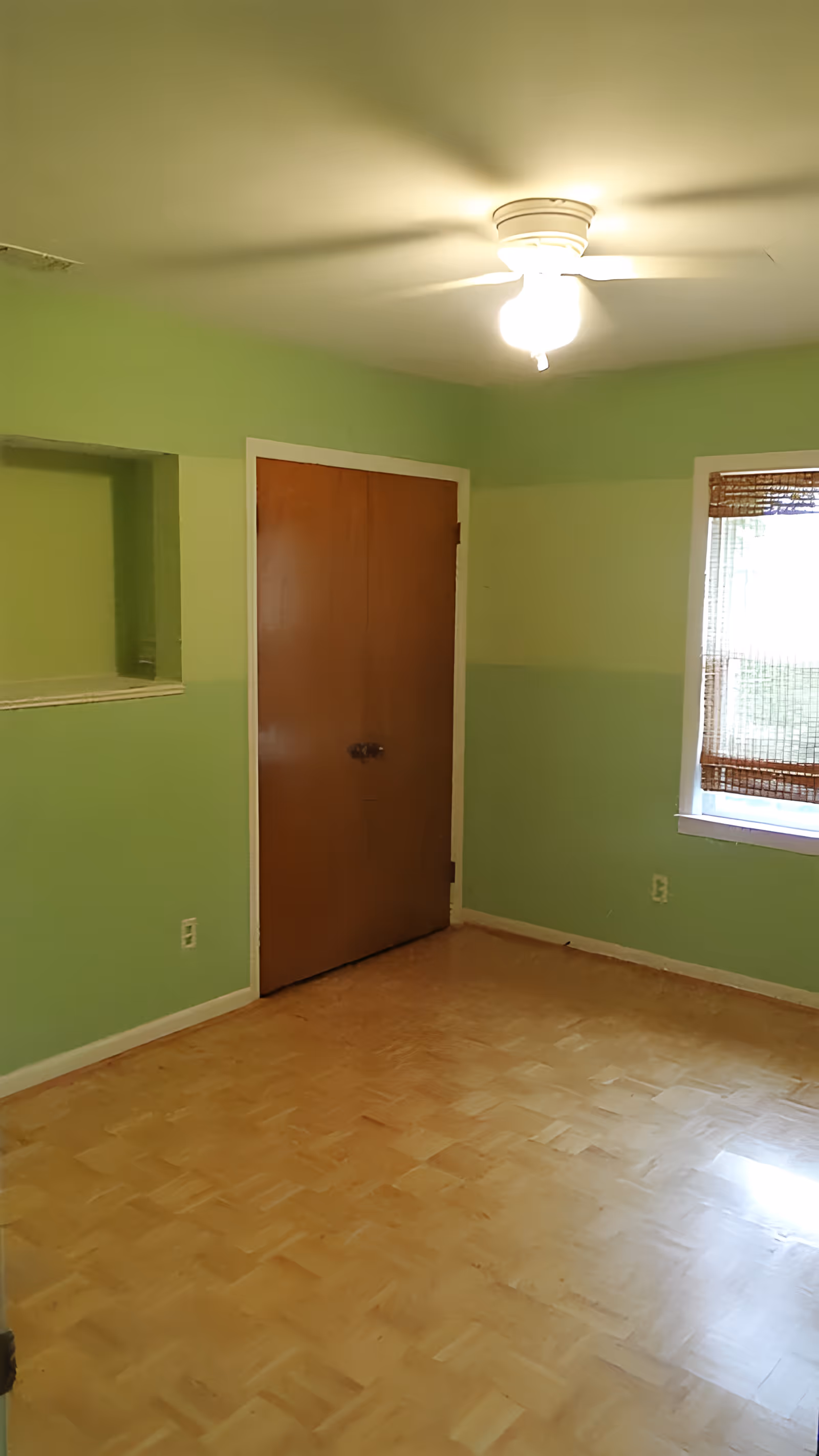 Empty room with two-tone light green walls, parquet floor, a closed wooden door, a window with a bamboo blind, and a ceiling light fixture.