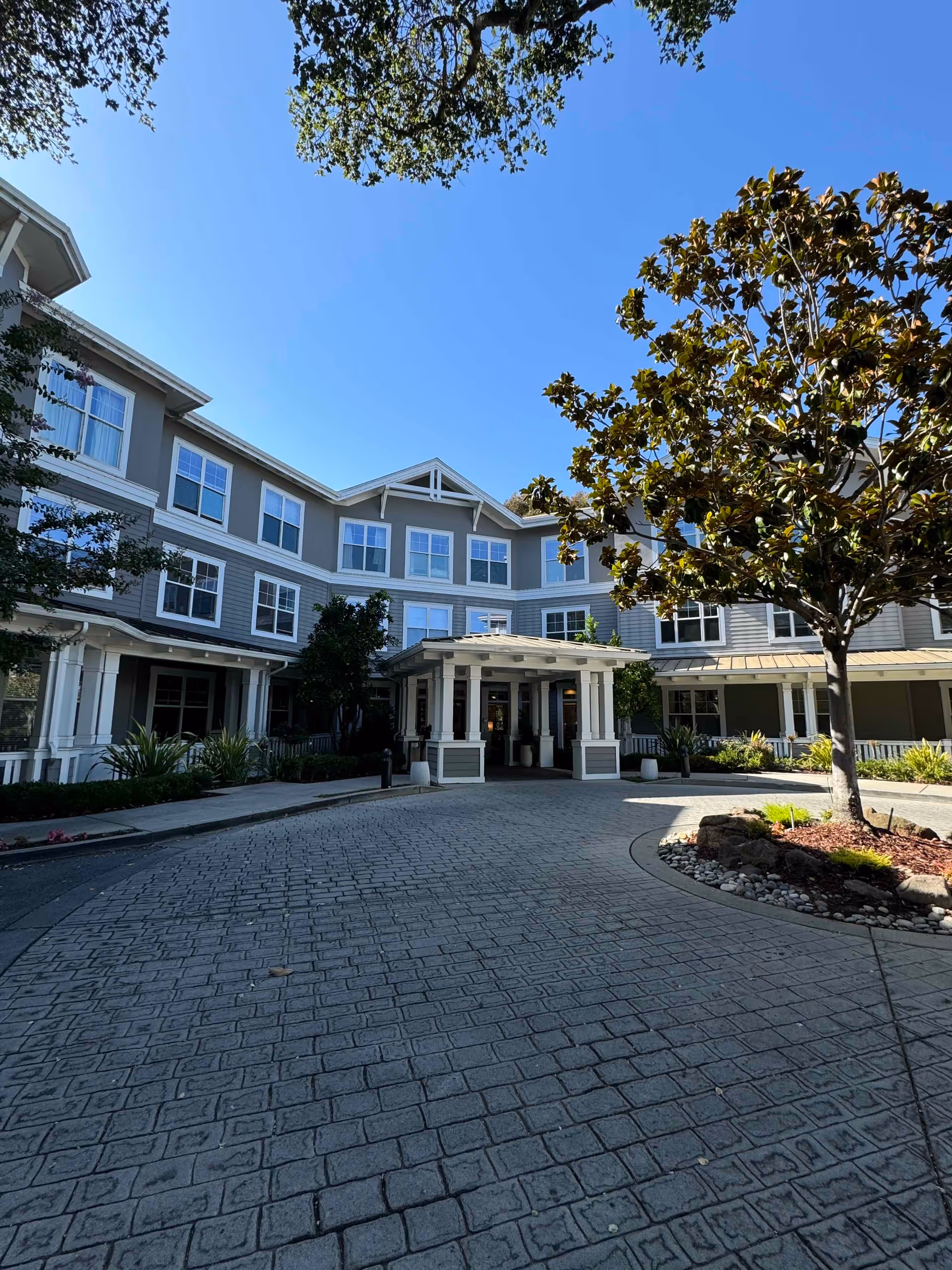 Front entrance of a multi-story senior living building with a covered porte-cochère, circular paved driveway, and landscaping.