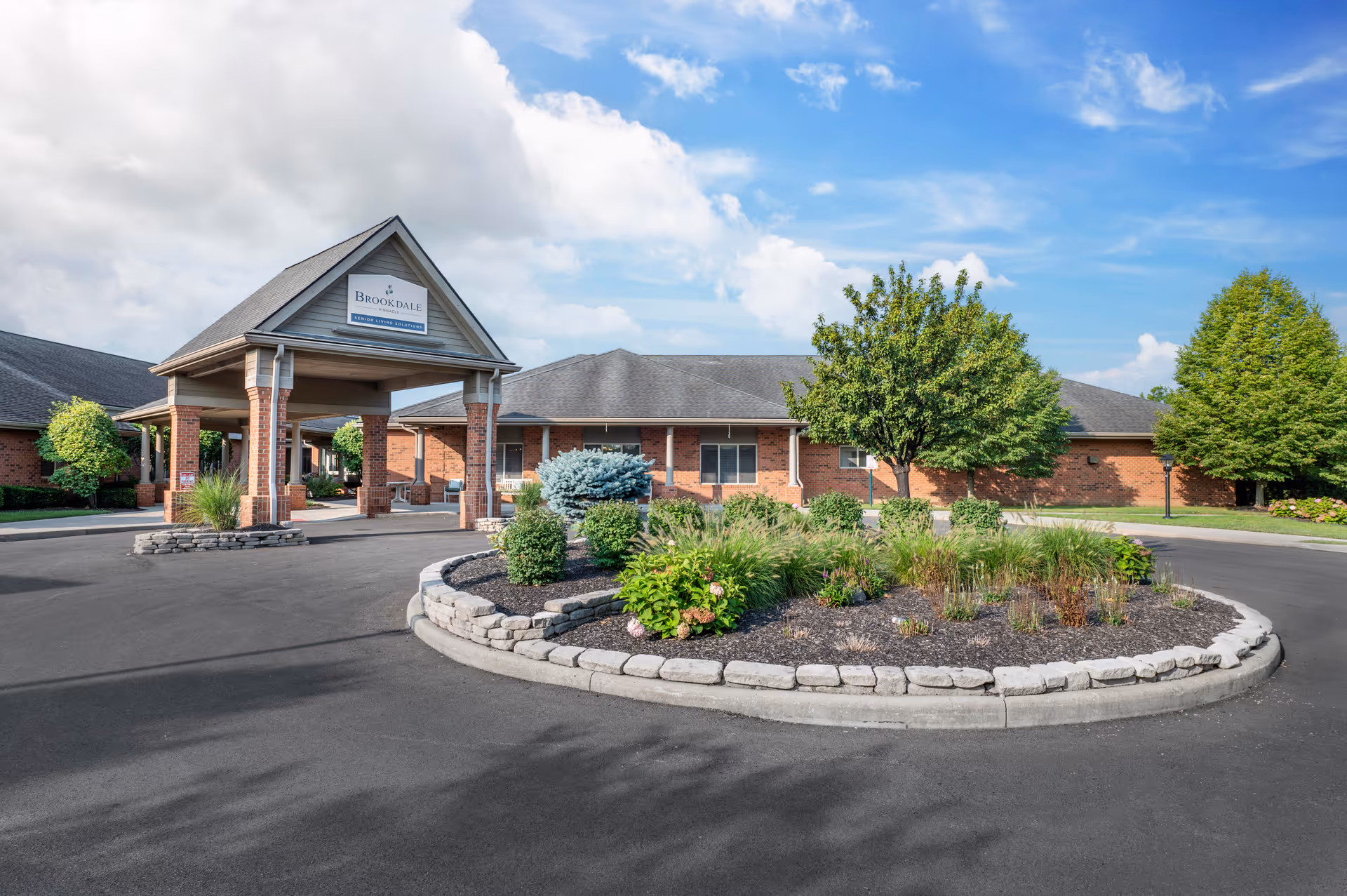 Exterior view of Brookdale Pinnacle senior living facility showing a circular driveway with landscaped center island, brick building with a covered entrance, and trees under a partly cloudy sky.