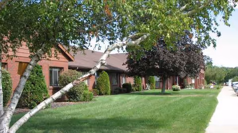 Oakbrook Village exterior: brick building with green lawn, trees, and a sidewalk.