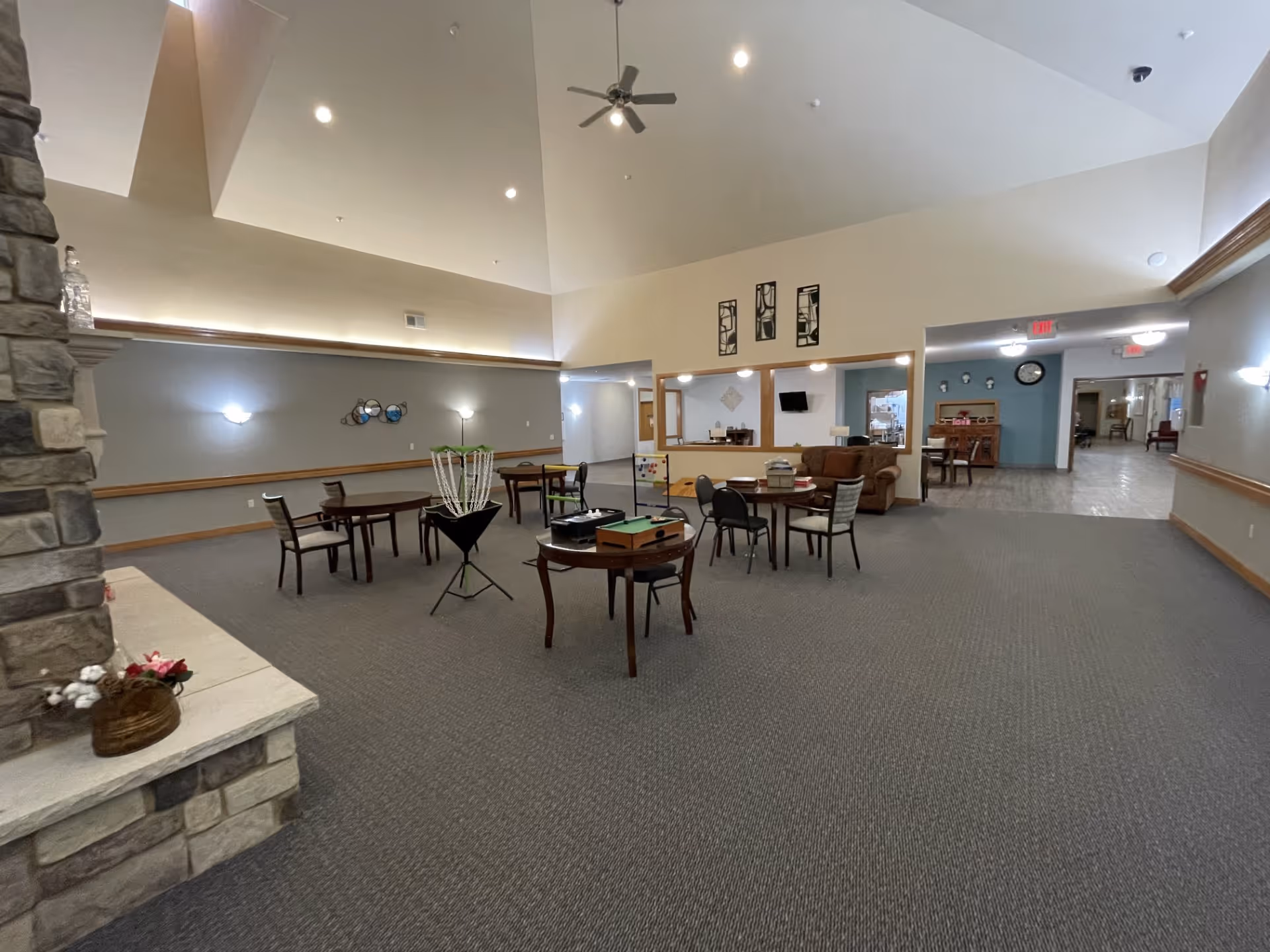 A spacious common area in a senior living facility with high vaulted ceilings and a ceiling fan. The room has several small tables and chairs arranged for activities, including a disc golf basket and various game boards. There is a stone fireplace on the left side with a small flower arrangement on the hearth. The walls are painted in neutral tones with soft lighting fixtures, and there is a seating area with a brown couch near an open window to another room. The far end of the room leads to a hallway with more seating and a clock on the wall.