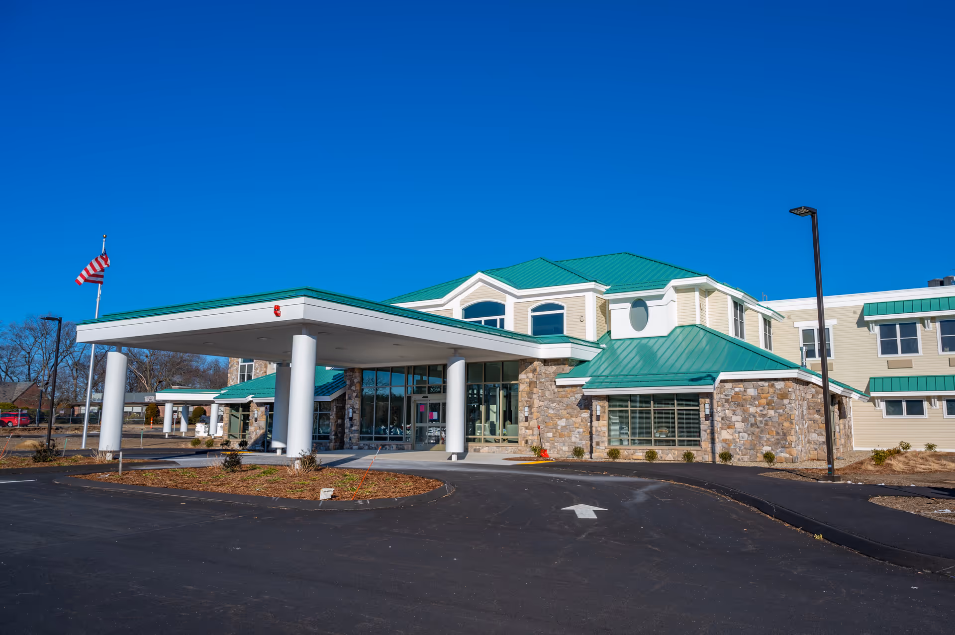 Exterior view of a modern skilled nursing center building with a green roof, stone and beige siding, a covered entrance supported by white columns, an American flag on a flagpole, and a clear blue sky.