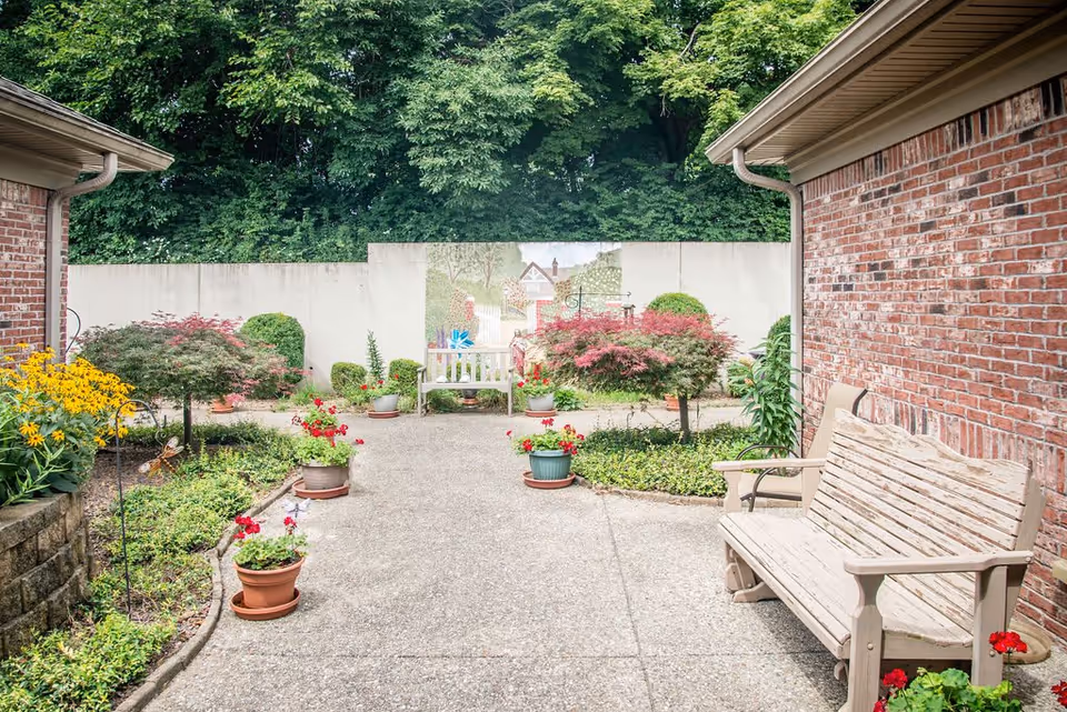 Outdoor courtyard area with a concrete pathway lined with potted plants and flowers. There are two wooden benches on either side, brick walls on the right and left, and a mural painted on a white wall at the far end. Green trees and bushes surround the area.