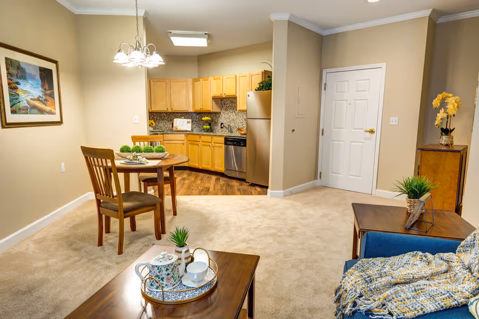 Interior view of a senior living facility apartment showing a small dining area with a round wooden table and two chairs, a kitchen with wooden cabinets and stainless steel appliances, and a living area with a blue couch covered with a knitted throw blanket. There is a coffee table with a tea set and a side table with a plant and picture frame. The walls are beige, and there is a framed painting on the wall near the dining area.