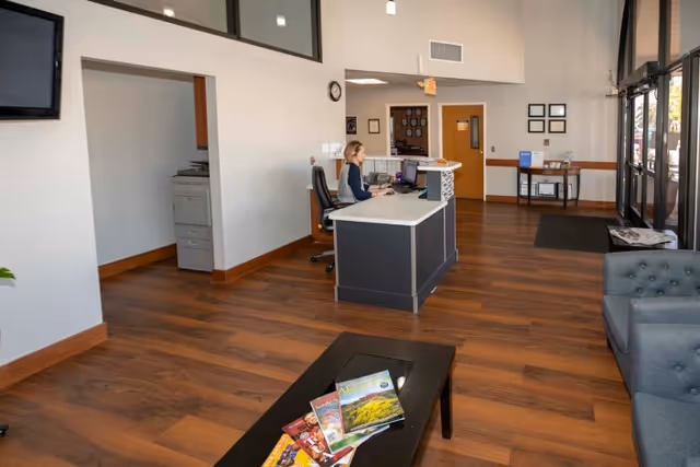 Reception lobby with a front desk where a staff member works, seating area, coffee table with magazines, and glass entrance doors.