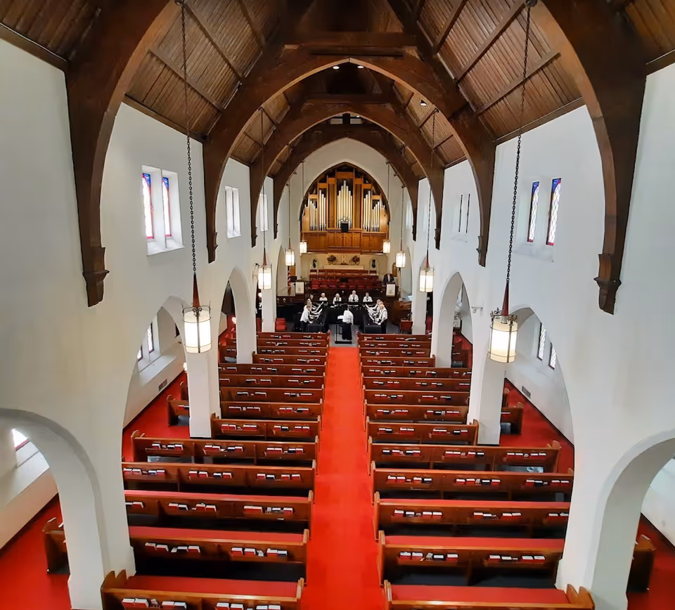 High-angle view down a church sanctuary interior with wooden pews, a red carpeted center aisle, arched wooden ceiling, and an organ and choir at the front.