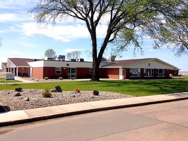 Exterior view of a single-story brick building with a large tree in front, green lawn, and a landscaped area with rocks and small shrubs under a partly cloudy sky.