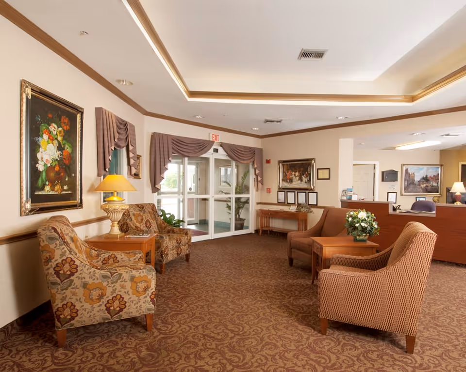 A cozy and well-lit senior living facility lobby with patterned carpet and comfortable upholstered chairs arranged around wooden side tables. The walls are decorated with framed paintings and draped curtains above glass double doors. A reception desk is visible in the background with additional artwork and a lamp.