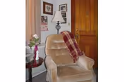 A cozy corner of a room featuring a beige upholstered armchair with a red and beige plaid throw draped over one arm. Next to the chair is a small round wooden side table with a vase holding a white flower. Behind the chair is a wooden door and a wall decorated with framed pictures and a table lamp with a white shade.
