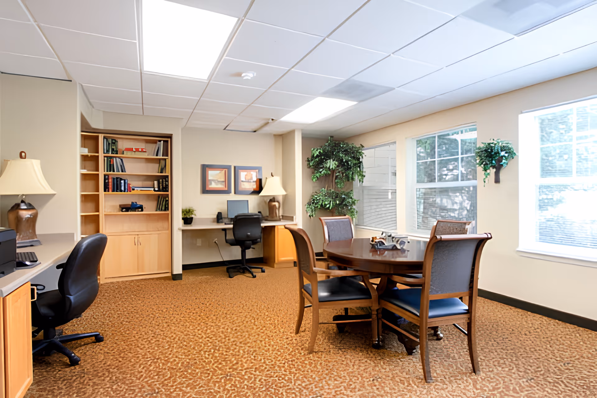A well-lit office or common area with a round wooden table surrounded by four chairs, two desks with office chairs, a computer, bookshelves, two table lamps, framed pictures on the wall, and large windows letting in natural light. There is also a potted artificial plant in the corner.
