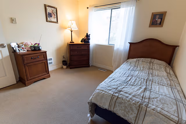 Small furnished bedroom with a single bed, wooden dressers, a lamp, and a window with sheer curtains.